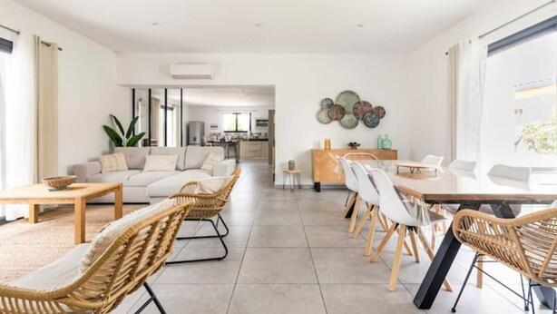 A spacious living area with a sofa, dining table, and chairs, a kitchen in the background, and a large window with curtains.
