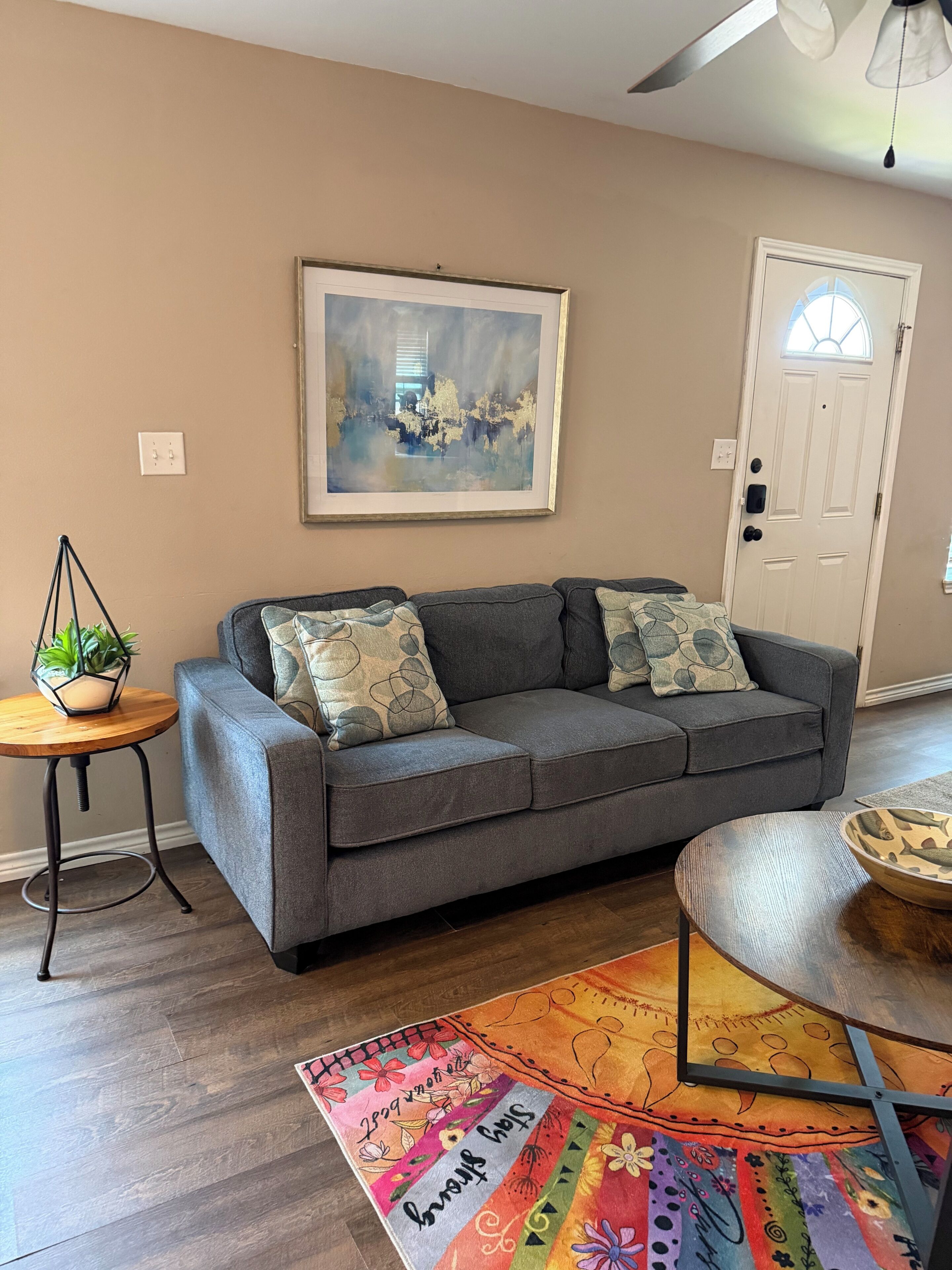 A living room with a gray sofa, a framed abstract painting, a small wooden table with a plant, and a colorful rug.