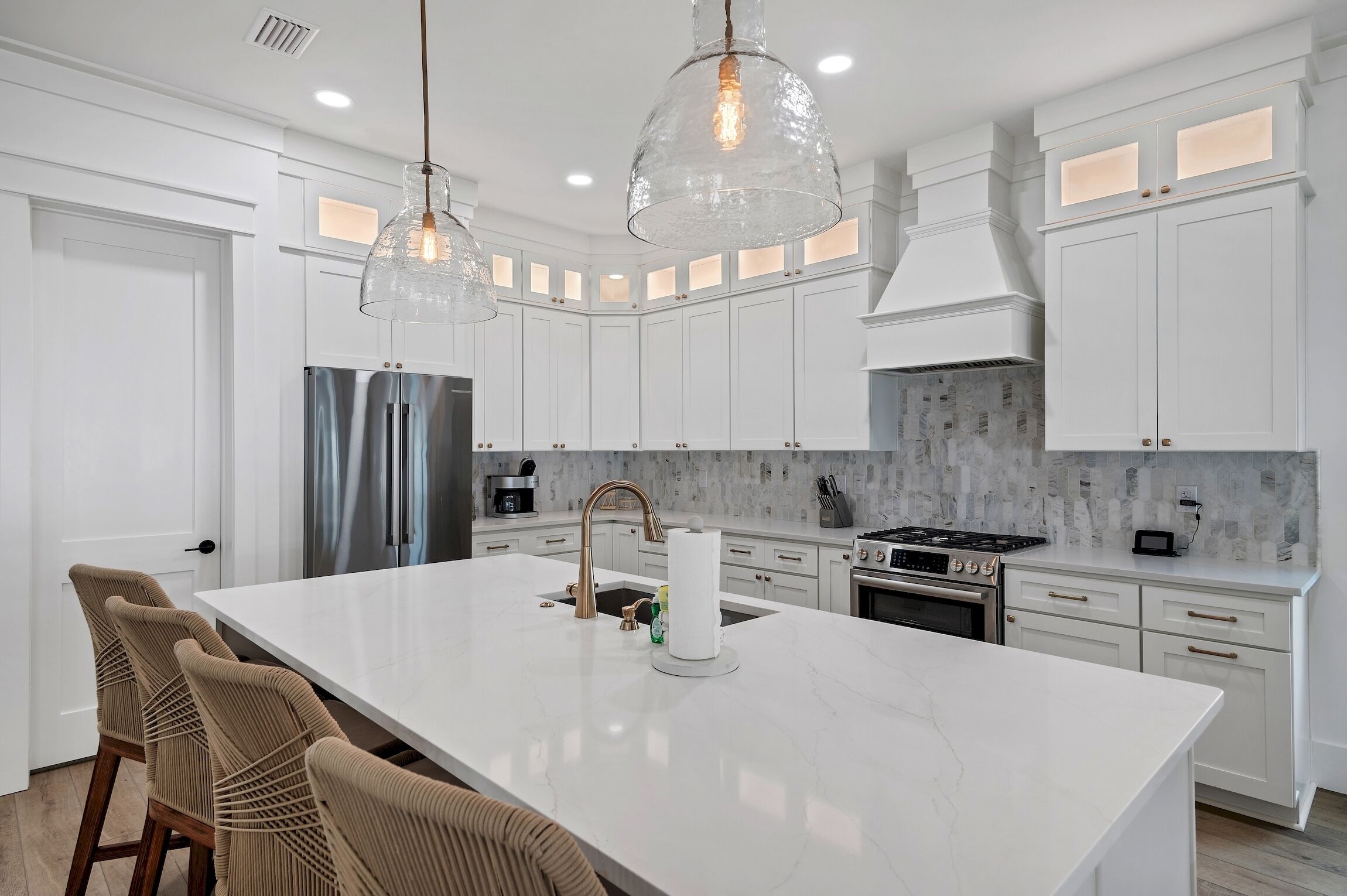 Une cuisine moderne avec des meubles blancs, un grand îlot central, des appareils en acier inoxydable et des luminaires suspendus.