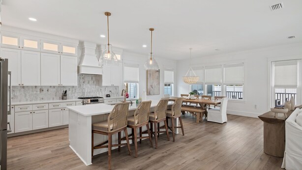 A modern kitchen with white cabinetry, a central island with bar stools, pendant lighting, and a view of the city through large windows. - Luxe 5BR with Pool, Hot Tub, Outdoor Kitchen, Bikes, Close to Beach & Seaside (Santa Rosa Beach)