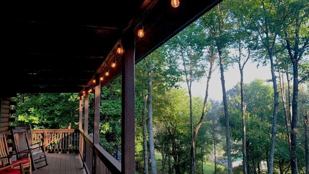 A wooden deck with hanging lights, wooden chairs, and a view of a forested area. - Cozy Mtn Log Cabin, wonderful views, 5 mins from Hayesville town sq, sleeps 8. (Hayesville)