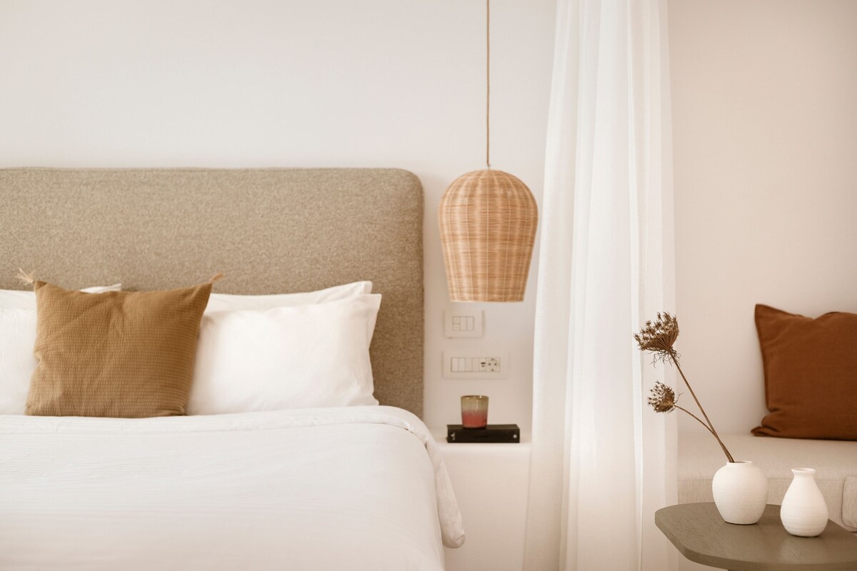 Quarto bem arrumado com cama, travesseiros, mesa de cabeceira com um vaso e abajur, e vista para o exterior.