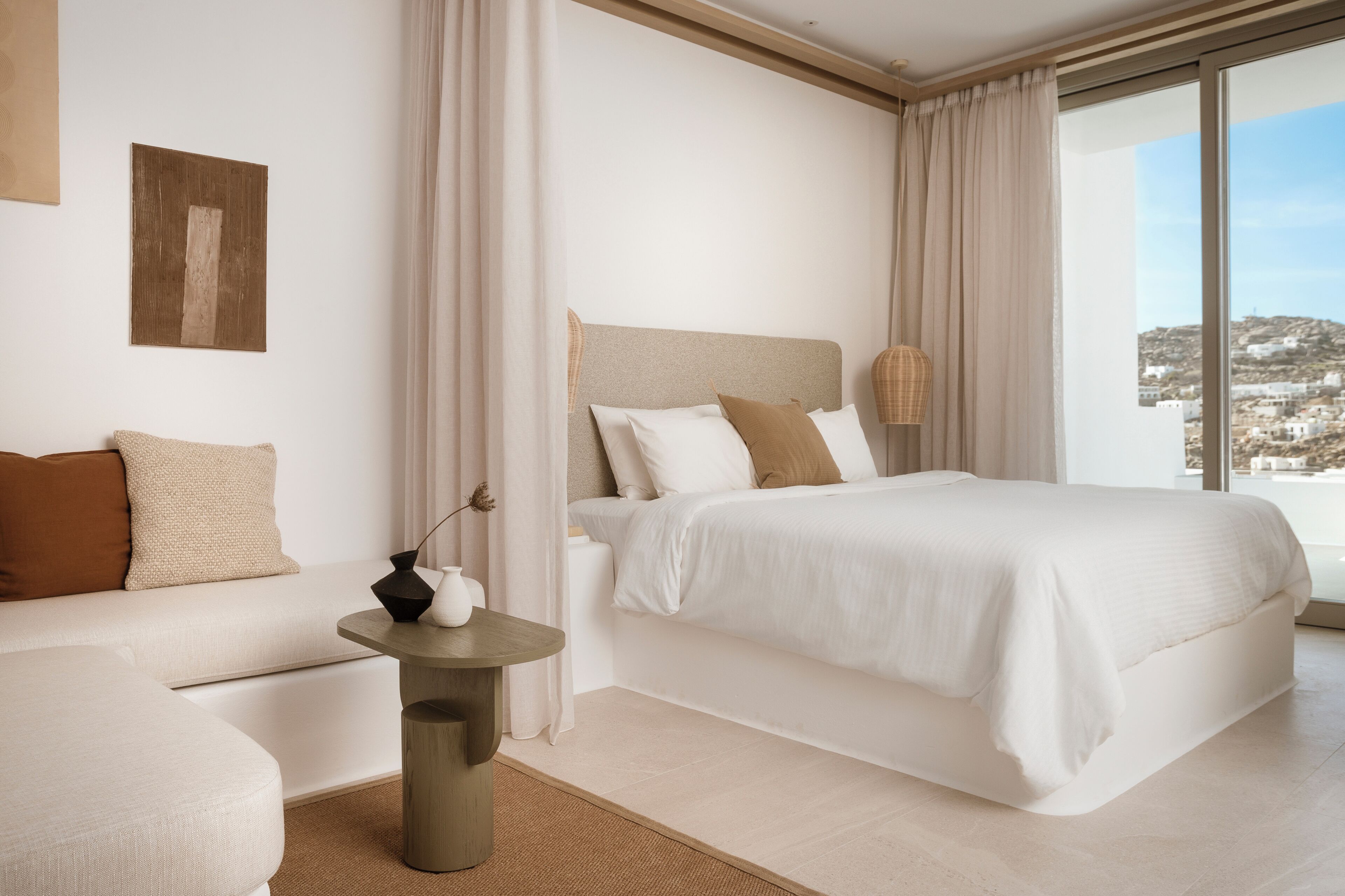 A modern hotel room with a large bed, a sofa, a small table, and a view of the city.