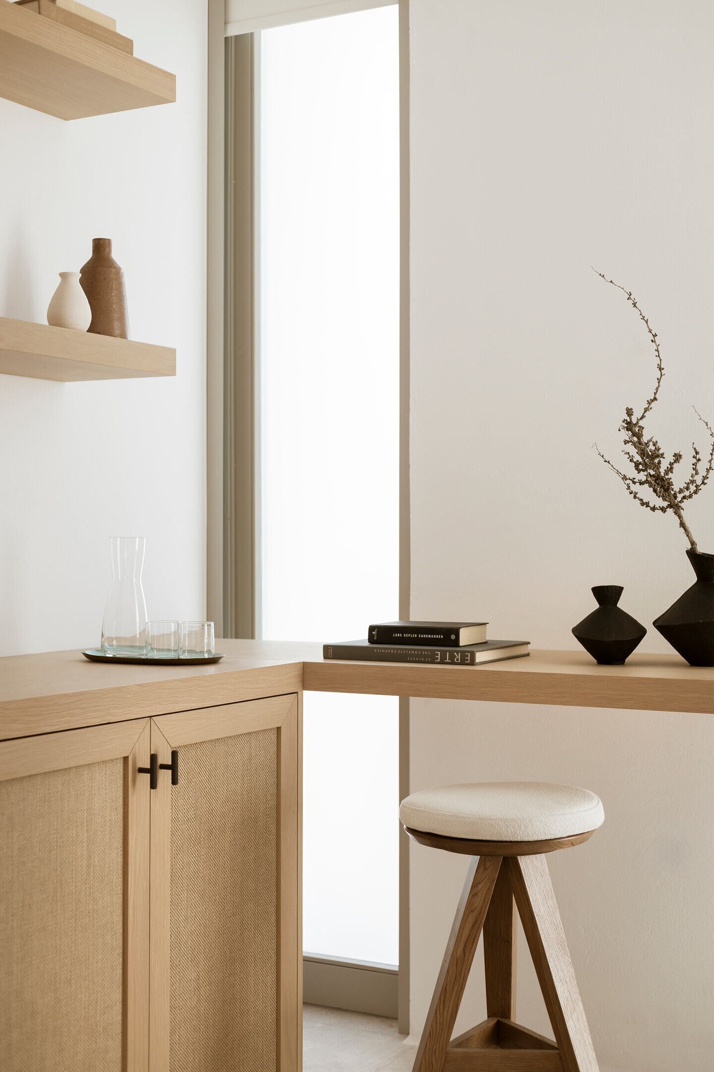 A minimalist room with a wooden desk, a stool, and a window with natural light.