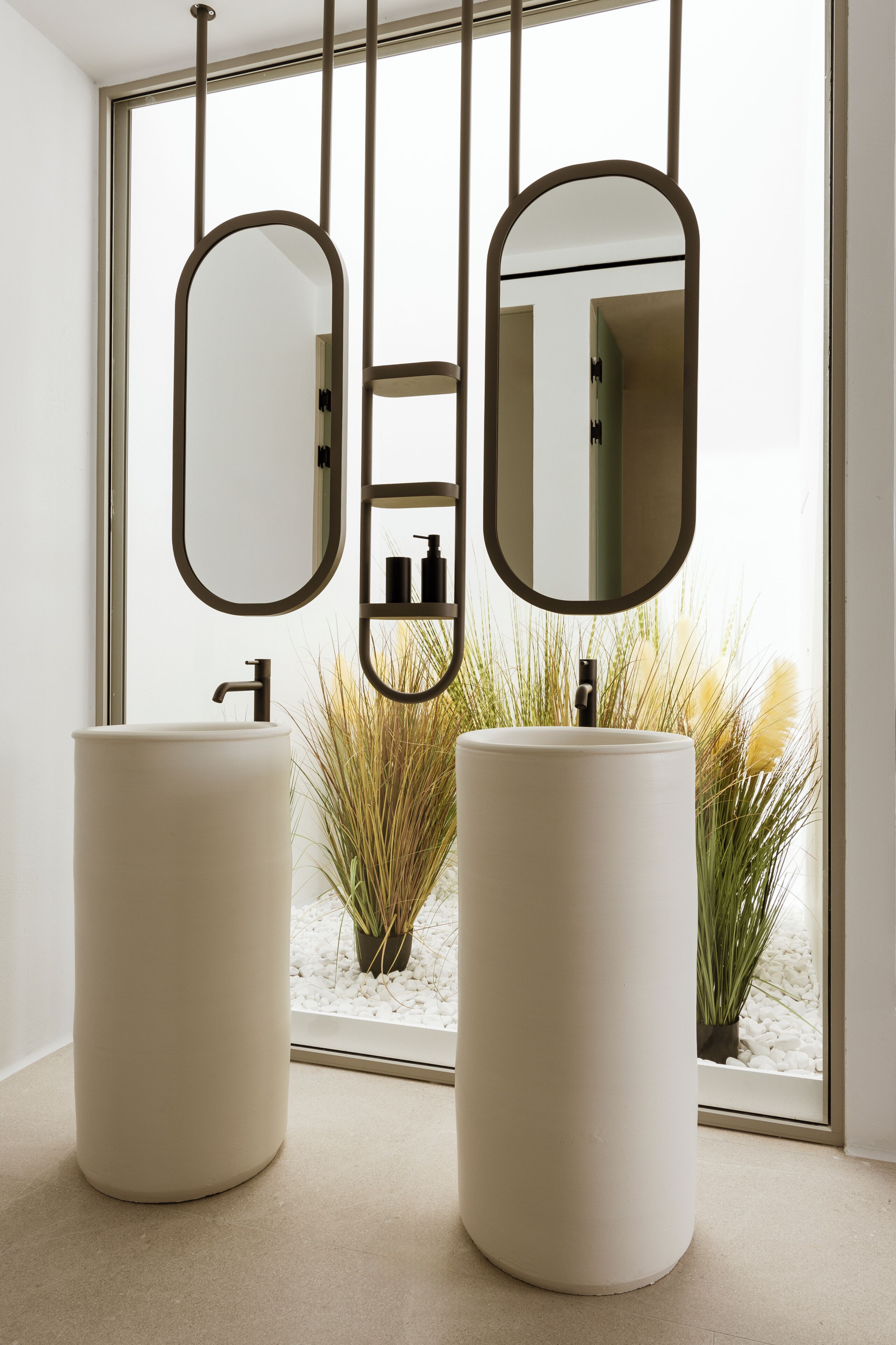 Une salle de bain moderne avec deux grands lavabos cylindriques, un miroir et une vue sur un jardin, grâce à une grande fenêtre.