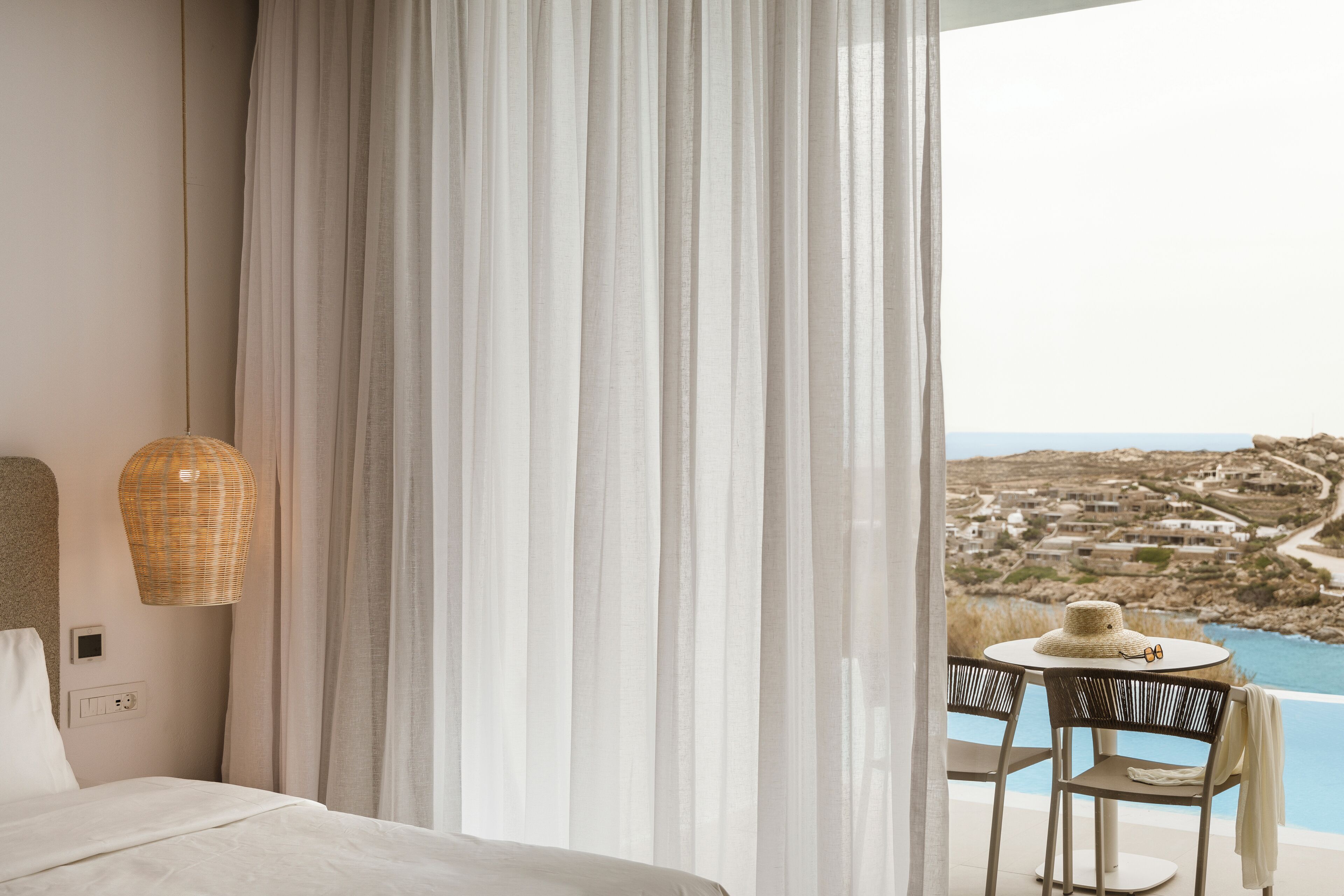 A bedroom with a bed, a table with chairs, a wicker lamp, and a view of a coastal landscape.