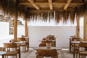 A restaurant with wooden tables and chairs, a thatched roof, and a view of the landscape.