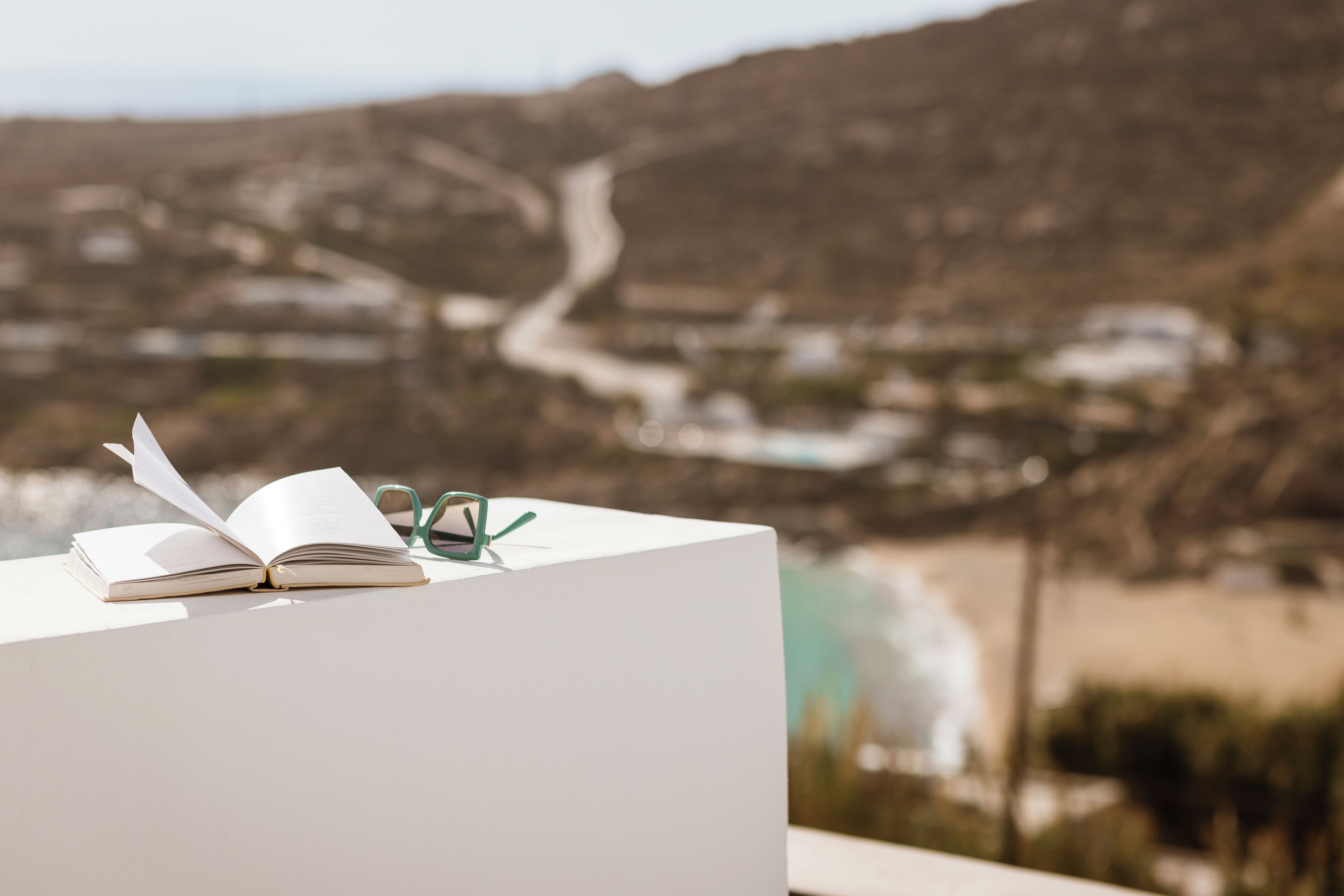 Un libro aperto su una superficie bianca, con un paio di occhiali accanto, con vista su un paesaggio costiero con edifici e una strada tortuosa.