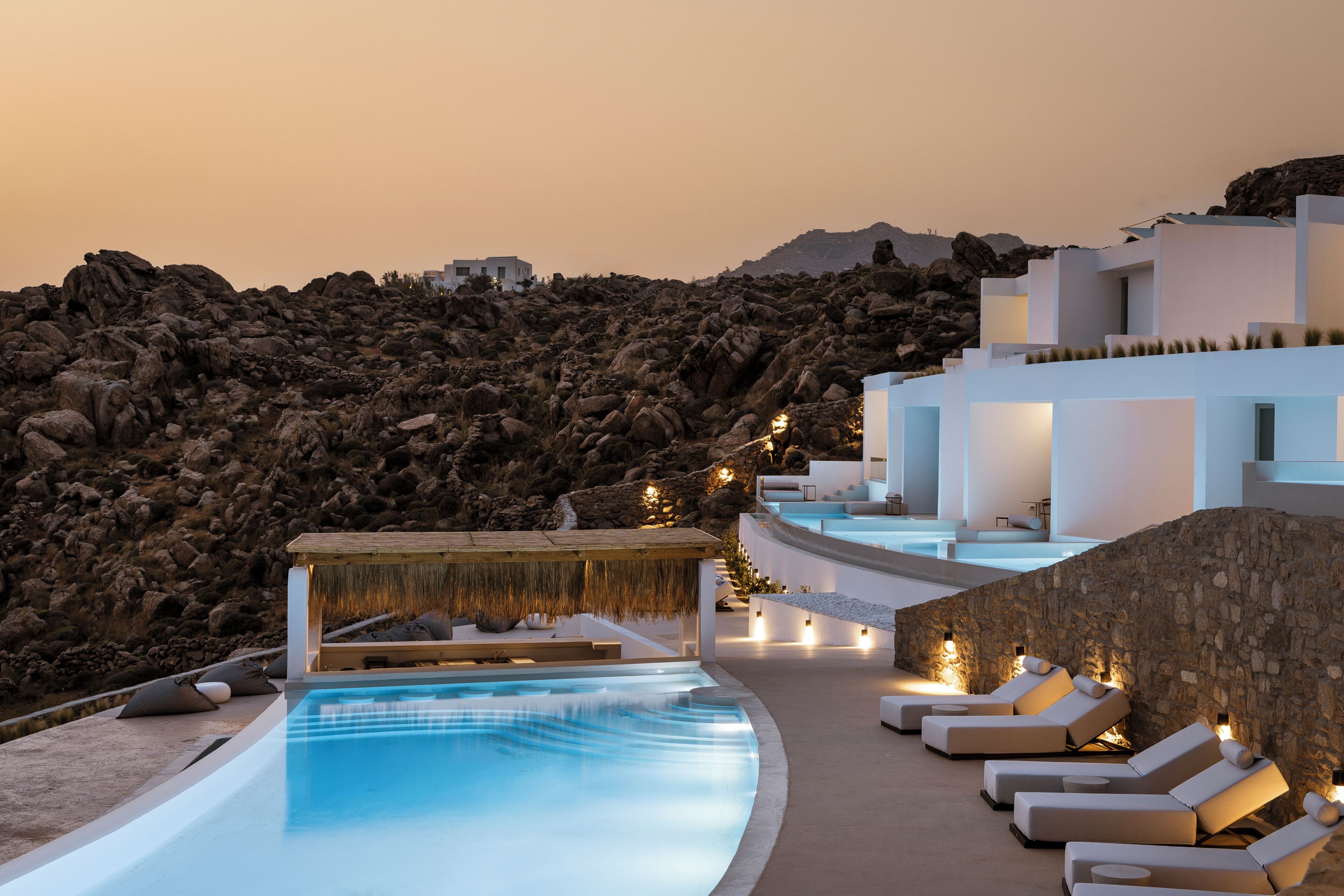 A poolside area with lounge chairs, a thatched-roof structure, and a view of rocky terrain.