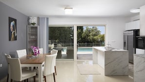 A modern kitchen with a marble island, a dining area with white chairs, and a glass door leading to a pool.