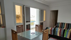 A dining area with a glass table and wooden chairs, a colorful sofa, and a view through a sliding door. - Cozi Coliving (Mezraia)
