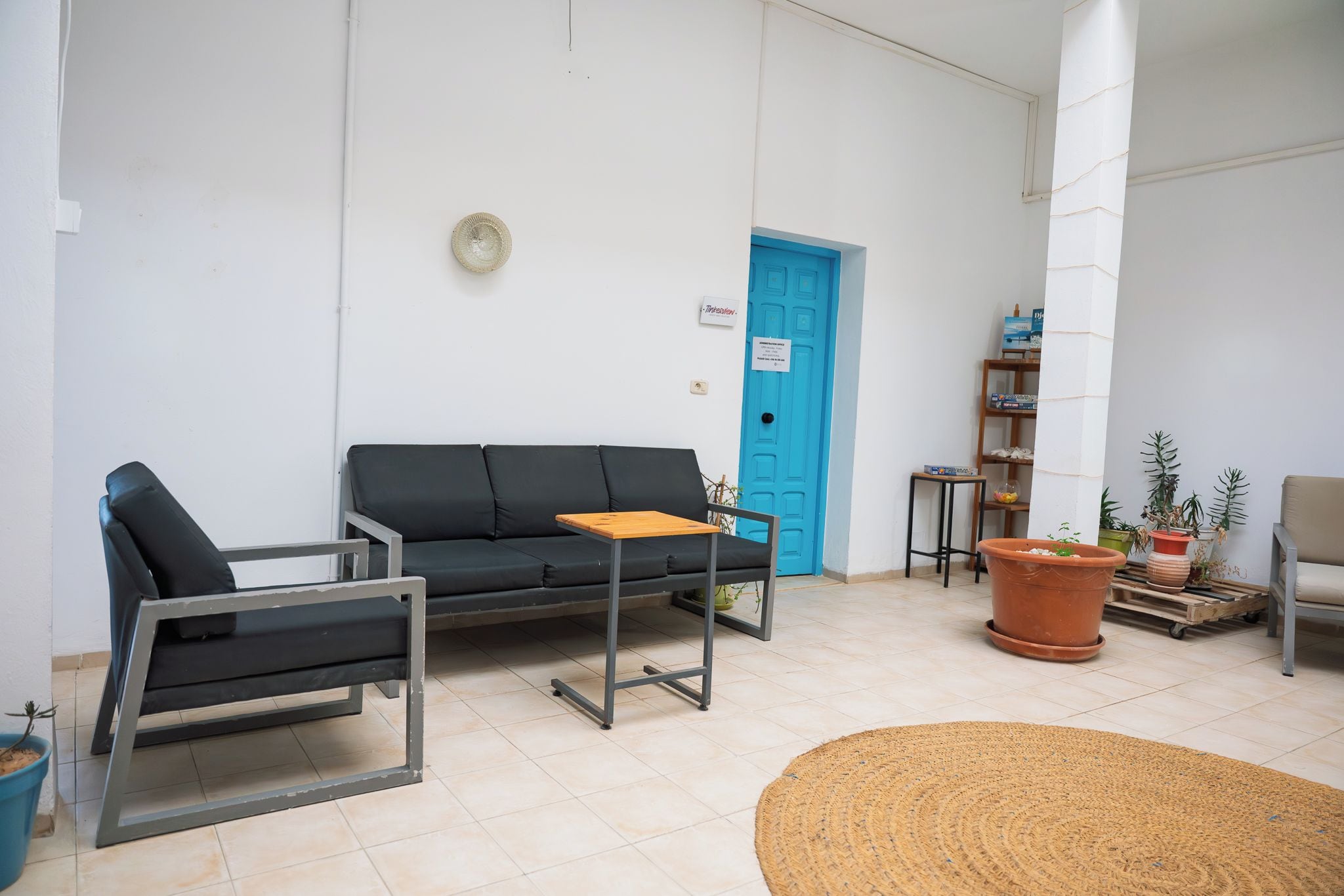 Una habitación con un sofá negro, una mesita, una puerta azul y una estantería con plantas.