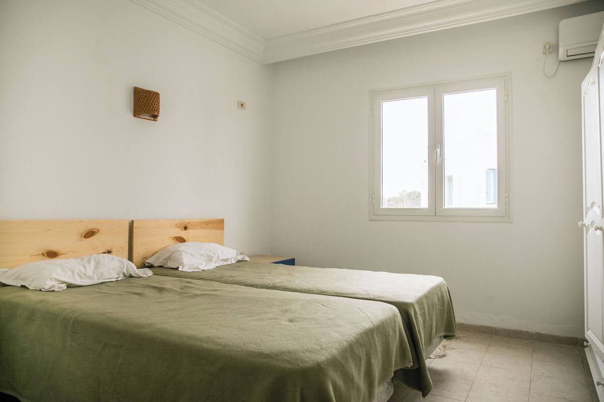 Une chambre avec deux lits, une tête de lit en bois, une fenêtre et un appareil de climatisation.