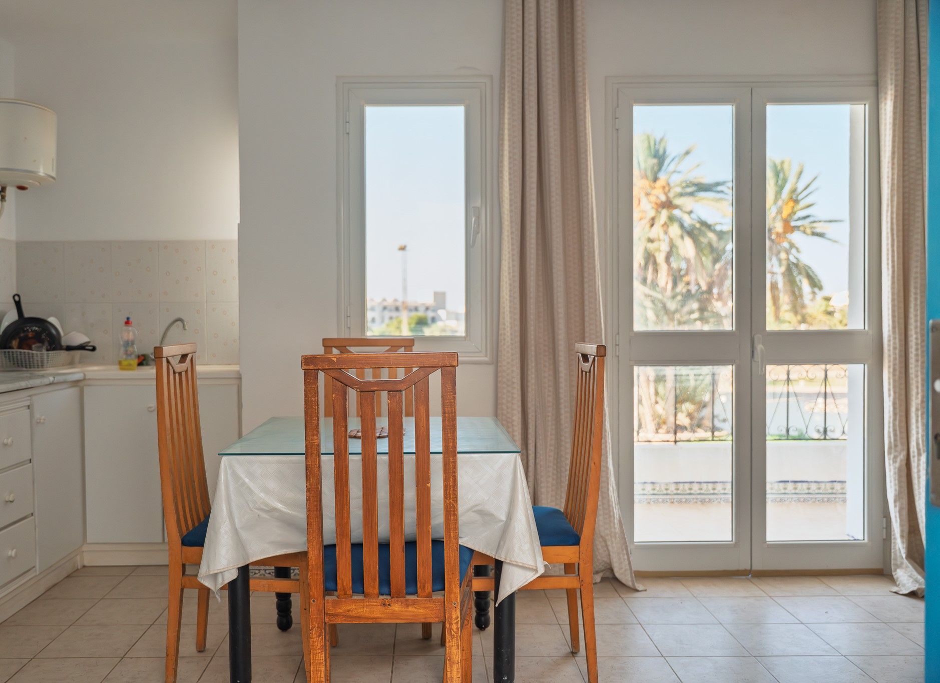 A dining area with a table and chairs, a kitchen area with a sink and utensils, and a view of palm trees through a door.