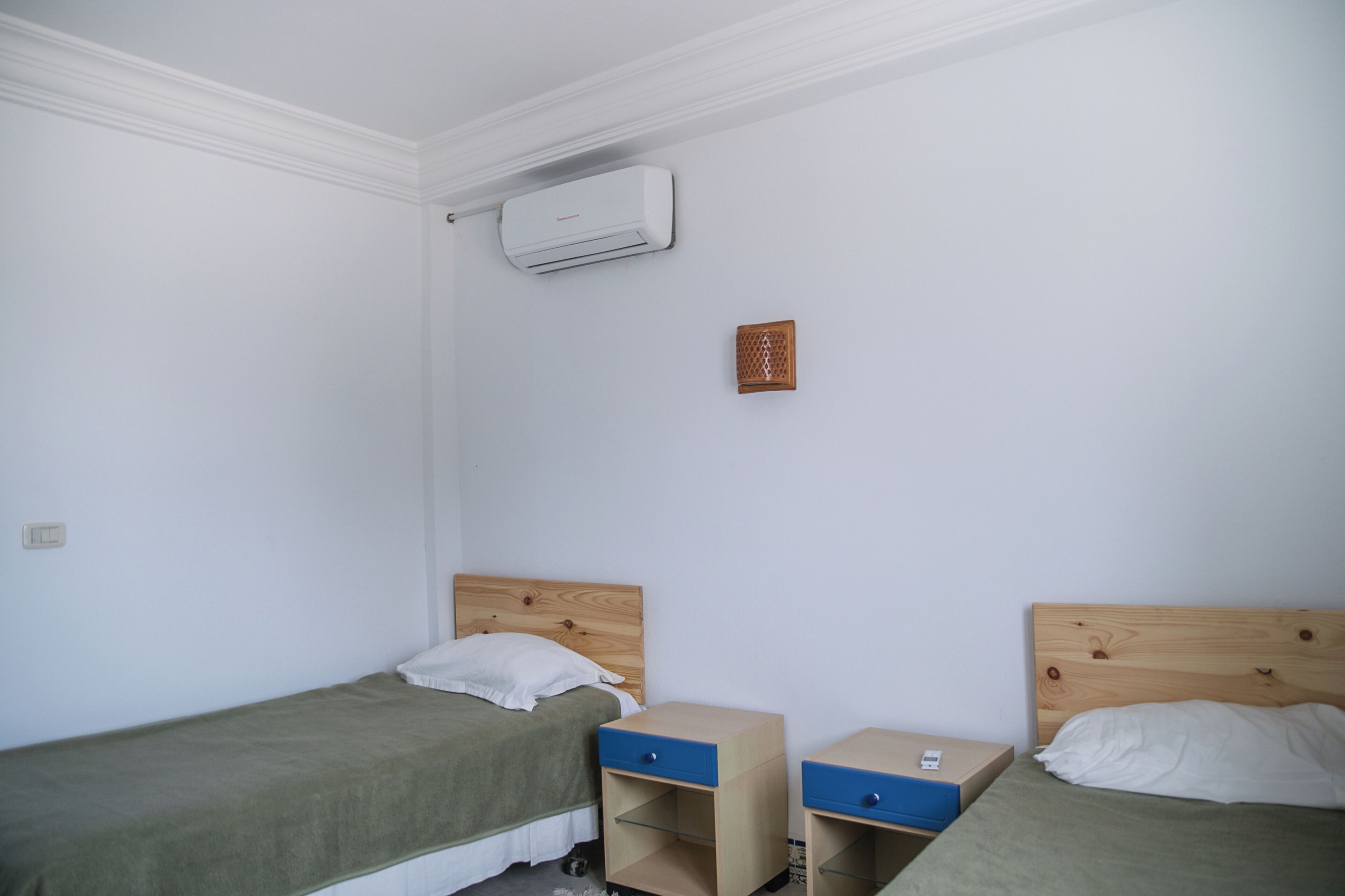 Une petite chambre propre, avec deux lits simples, une tête de lit en bois, une petite table de chevet avec un tiroir et un climatiseur fixé au mur.