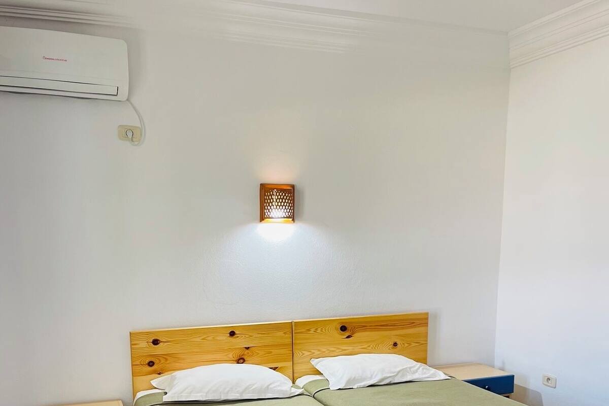 Un lit double avec une literie verte, une tête de lit en bois, une table de chevet avec un tiroir, un luminaire fixé au mur et un appareil de climatisation.