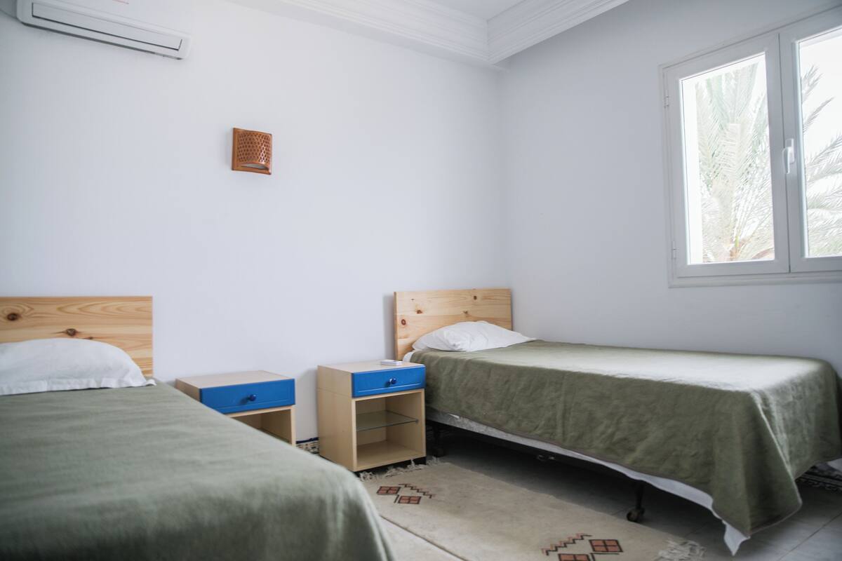Une chambre avec deux lits simples, une petite table de chevet en bois avec un tiroir bleu, un appareil de climatisation blanc fixé au mur et une fenêtre donnant sur des palmiers.