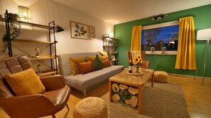 A modern living room with a sofa, armchair, and coffee table, a bookshelf, and a window with curtains. - Plant Apartment - Large balcony with barbecue (Eutingen im Gäu)