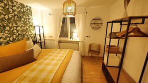 A bedroom with a bed, a chair, a wardrobe, a window with curtains, a wall-mounted shelf, and a hanging light fixture. - Plant Apartment - Large balcony with barbecue (Eutingen im Gäu)
