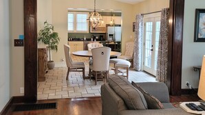 A spacious living room with a gray sofa, wooden flooring, and a ceiling fan. A dining area with a table and chairs is visible through an open doorway.