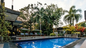 A swimming pool with lounge chairs and a covered seating area, surrounded by tropical plants and palm trees.