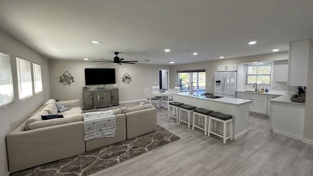 A modern living room with a sofa, a TV, and a ceiling fan. There is a kitchen area with a bar stool and a window. - 4 bed/ 3 bath Villa Heated Pool (Cape Coral)