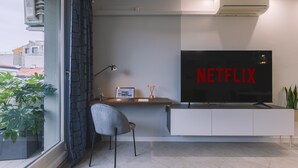 Una sala de estar moderna con un televisor de pantalla plana grande montado sobre un mueble blanco, un escritorio con una computadora portátil, una silla gris y una planta en maceta.