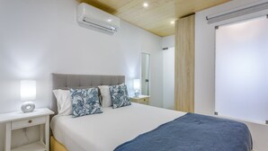 A bedroom with a bed, bedside tables, a lamp, an air conditioner, a mirror, and a window with blinds.