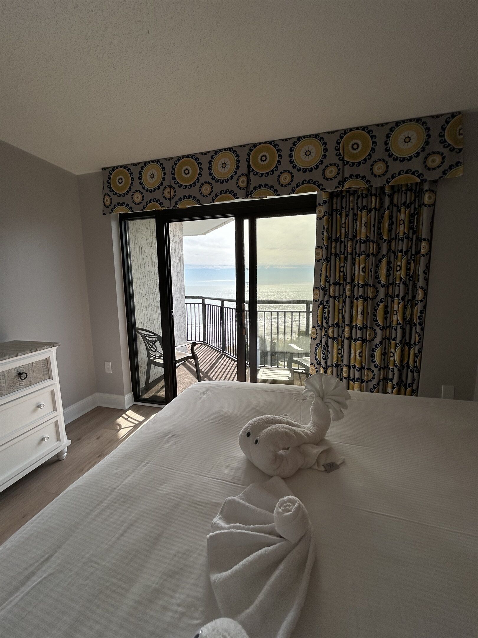 A hotel room with a large bed, a dresser, and a view of the beach through a sliding glass door.