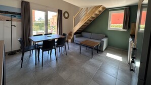 A modern living space with a dining area, a sofa, a staircase, and a kitchen. - Mini Villa T3 de 48m2 (Lecci)