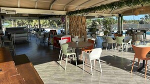 An outdoor cafe with tables and chairs, a pool in the background, and a covered area with a curved ceiling. - Mini Villa T3 de 48m2 (Lecci)