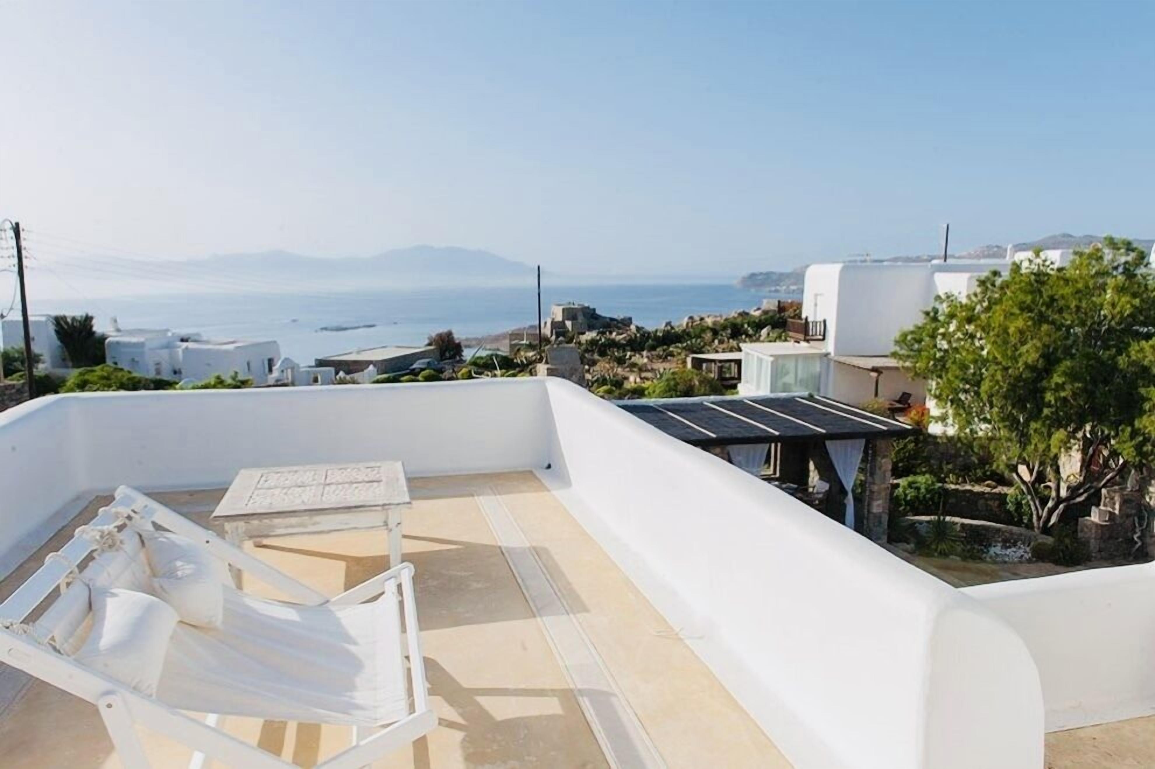 Eine Dachterrasse mit weißem Liegestuhl, einem kleinen Tisch und Blick auf das Meer sowie entfernte Berge.