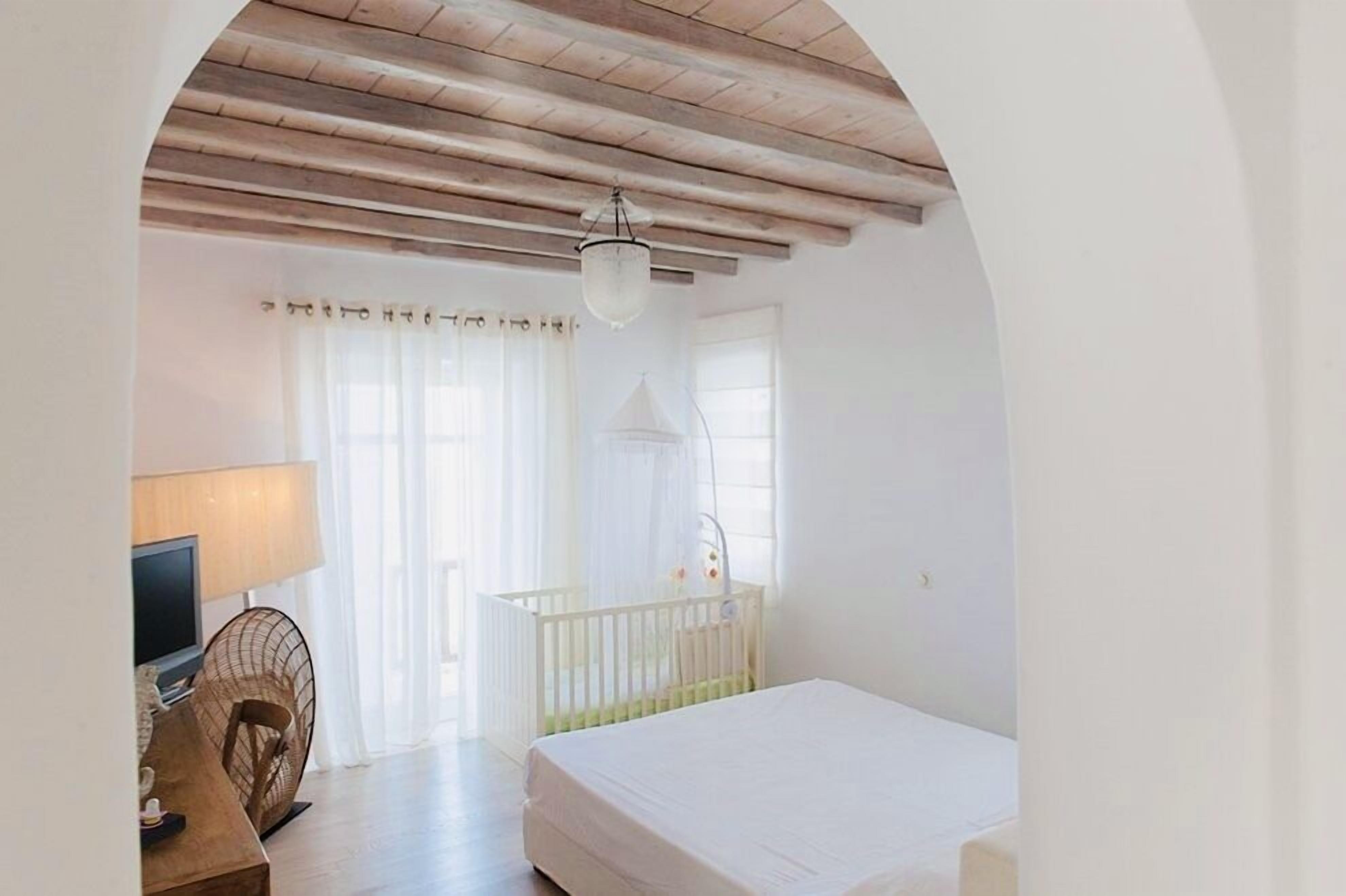 A bedroom with a wooden ceiling, a bed, a crib, a desk with a lamp, and a TV.