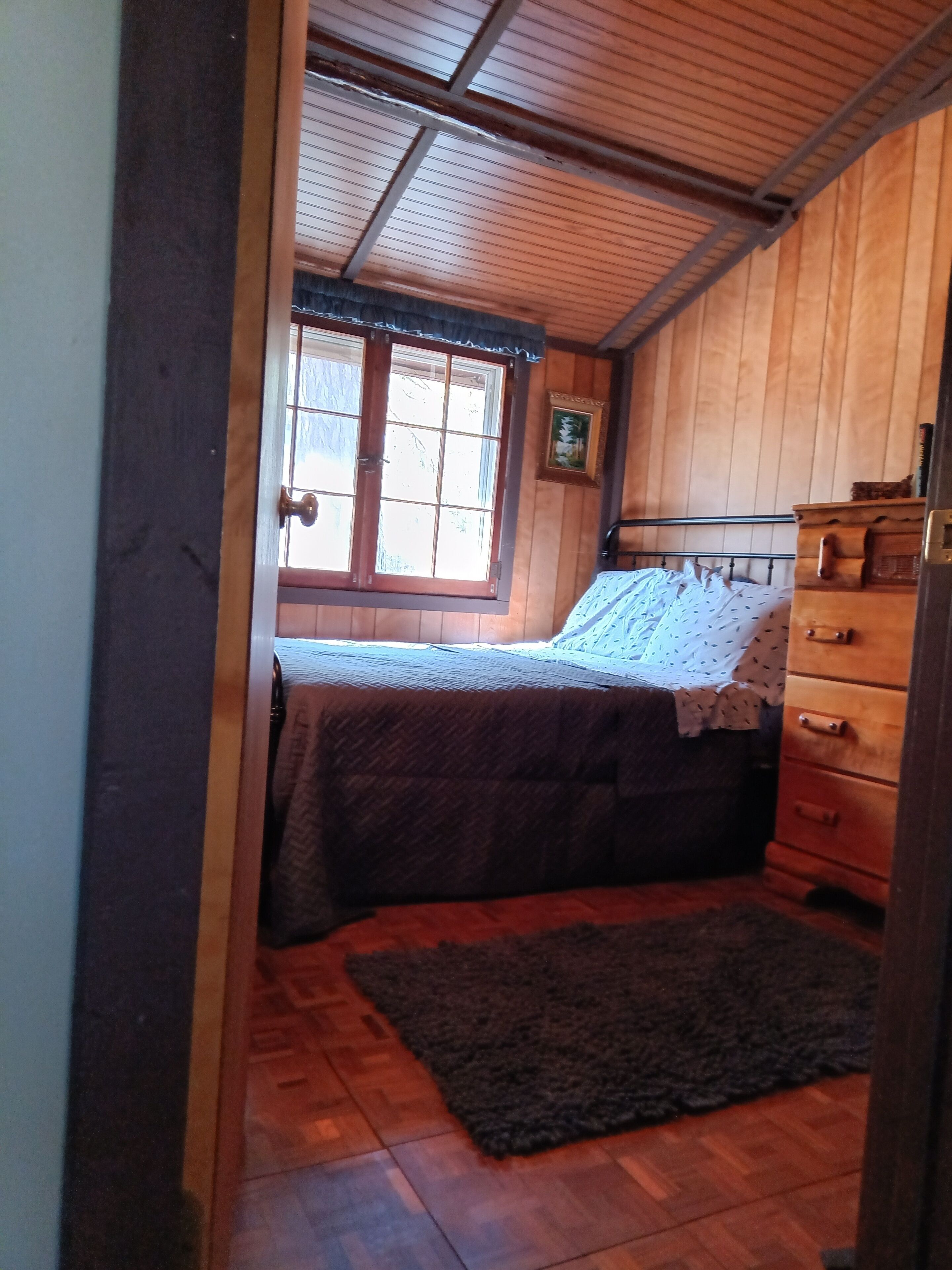A bedroom with a wooden ceiling, a window with a view, a bed with a patterned bedspread, a dresser, and a rug on the floor.