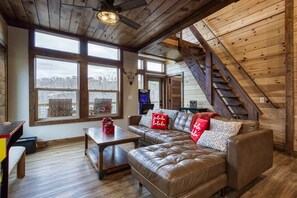 A cozy loft-style room with a leather sofa, wooden beams, and large windows offering a view of the outdoors. - Brand New Modern Mountain Chalet (Gatlinburg)