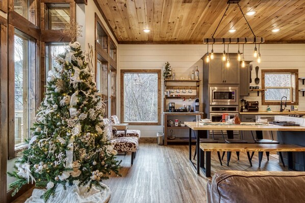 A cozy cabin interior with a large Christmas tree, wooden furniture, and a modern kitchen.