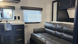 Ein kompaktes Wohnmobil-Interieur mit einer Küchenzeile, einem Ledersofa und einem Fenster mit Jalousien.