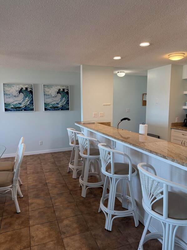 Une cuisine avec un coin bar, des tabourets de bar blancs, deux tableaux accrochés au mur et un sol carrelé.
