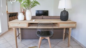 A modern living room with a wooden desk, a clear plastic chair, a brown sofa, a TV, a lamp, and a plant. - La Vue du Phare, Vue Mer Incroyable et Acces Privée Crique (Le Gosier)