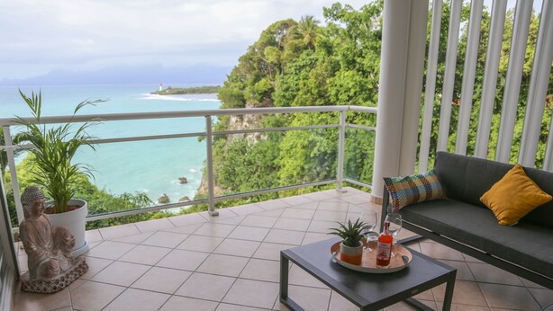 A balcony with a view of the ocean, a sofa with cushions, a small table with a plant and a bottle, and a Buddha statue. - La Vue du Phare, Vue Mer Incroyable et Acces Privée Crique (Le Gosier)