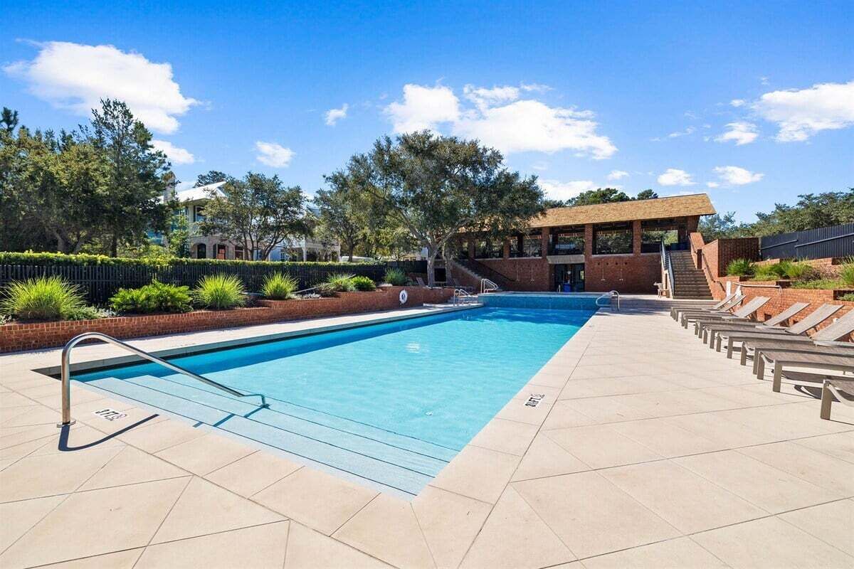 A rectangular swimming pool with a clear blue water, surrounded by a tiled deck. There are lounge chairs aligned along the poolside, and a covered seating area with a staircase leading to an upper level.