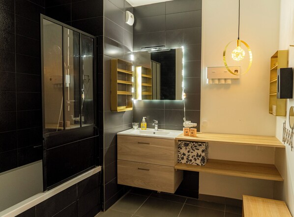 Un baño moderno con una pared de azulejos oscuros, cabina de ducha de vidrio, tocador de madera con espejo, y lámpara sobre el lavamanos.