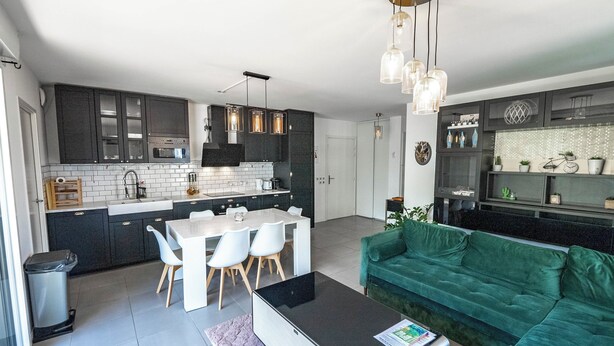 A modern kitchen with dark cabinets, a white countertop, and a dining area with a table and chairs. - Bright Apartment Near the lake (Bordeaux)