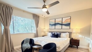 Ein modernes Schlafzimmer mit einem großen Bett, einem Deckenventilator, Blick auf die Stadt durch das Fenster und einem Nachttisch mit Lampe.