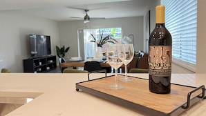 Eine Flasche Wein auf einem Tablett mit zwei Gläsern, ein Esstisch, Stühle, ein Fernseher und ein Deckenventilator in einem modernen Wohnzimmer.