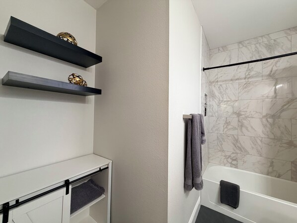Un baño moderno con una bañera blanca, azulejos de mármol y dos estantes negros con objetos decorativos.