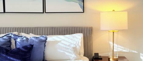 A neatly made bed with white linens and blue pillows, a bedside table with a lamp, and two abstract paintings on the wall.