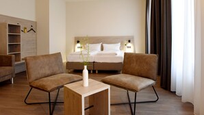 Habitación de hotel con cama, dos sillones, una mesita, un florero con planta y estante de madera.