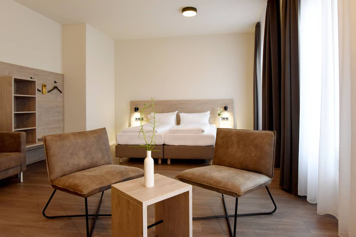 A hotel room with a bed, two armchairs, a small table, a vase with a plant, and a wooden shelf.