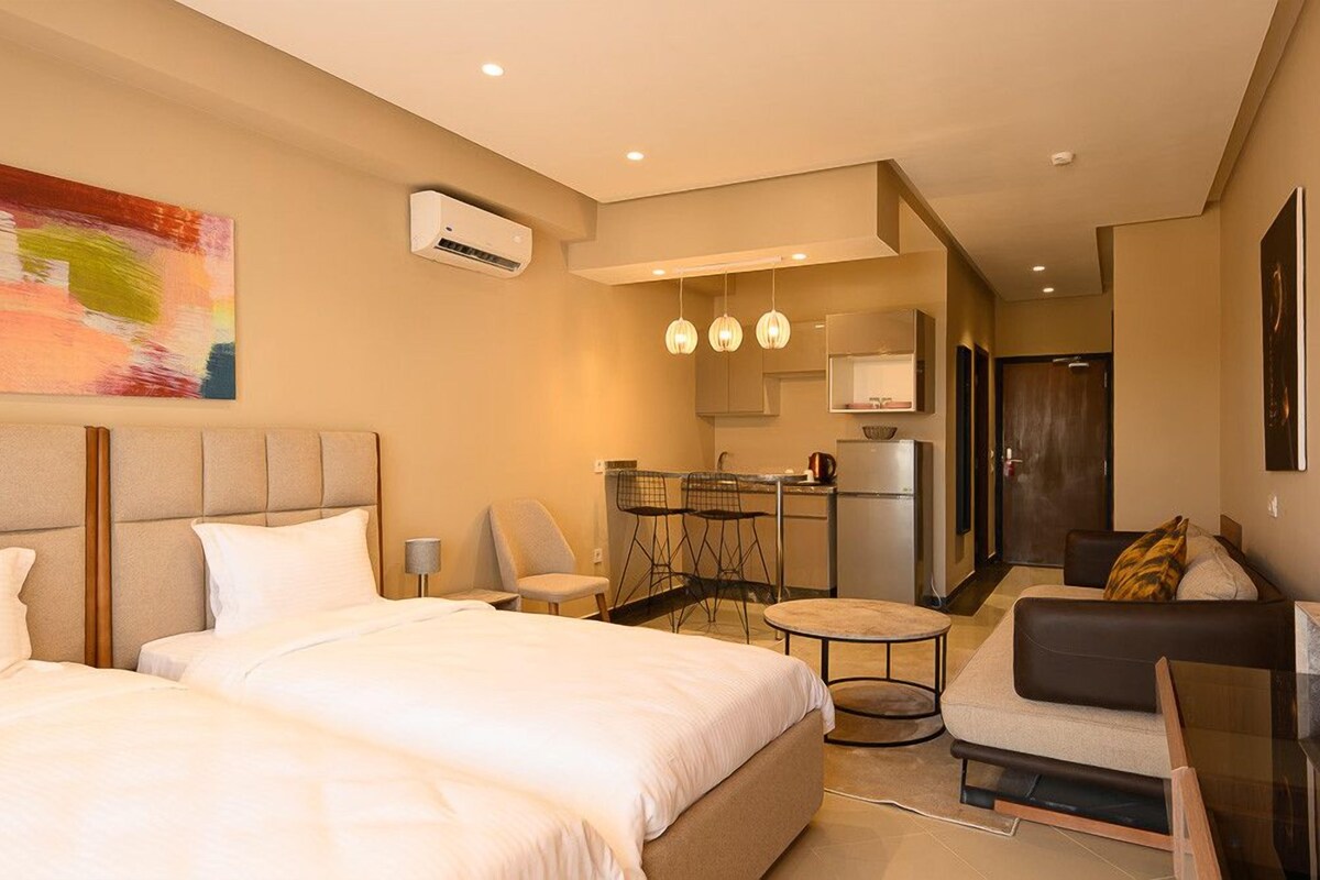 A hotel room with a large bed, a sofa, a small table, and a kitchenette area.